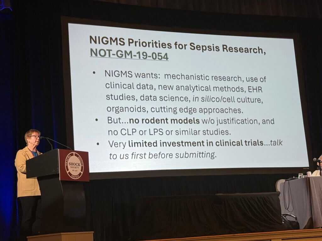 Dr. Rochelle Long stands at a podium next to a slide describing NIGMS priorities for sepsis research, which are focused on human sepsis research and not rodent models.